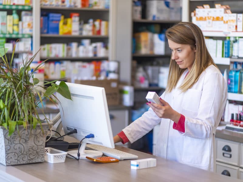 Female pharmacist working in chemist shop or pharmacy. Pharmacist using the computer at the pharmacy. Portrait of young female pharmacist holding medication while using computer at pharmacy counter